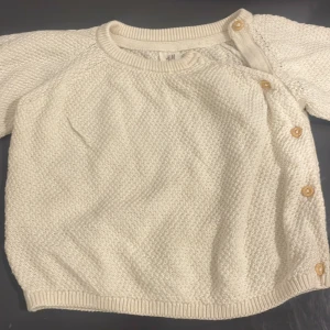 Beige stickad tröja från H&M med knappar - Superfin beige stickad tröja från H&M i ekologisk bomull. Tröjan har lång ärm, rund halsringning och snygga träknappar längs ena axeln och sidan för en cool detalj. Perfekt för lager-på-lager och enkel att matcha med allt.
