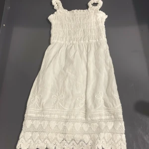Vit somrig klänning från TUMI - Supersöt vit klänning från TUMI med smock upptill och breda axelband. Klänningen har broderade blommor och hjärtan längs fållen samt spetsdetaljer. Perfekt för varma dagar och har en luftig känsla tack vare bomullsmaterialet. (till barn i ålder 2/3)