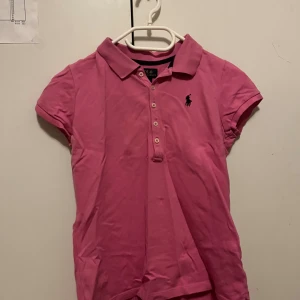 Rosa pikétröja från Ralph Lauren - Säljer en klassisk rosa pikétröja från Polo Ralph Lauren med korta ärmar och knappar framtill. Tröjan har den ikoniska broderade loggan på bröstet och är tillverkad i mjuk bomull. Perfekt för en sportig och stilren look.