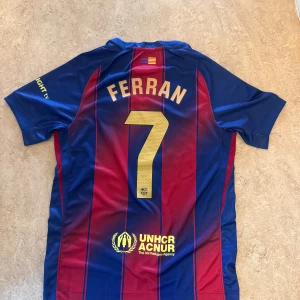 FC Barcelona Ferran #7 matchtröja Nike - Säljer en officiell FC Barcelona matchtröja från Nike med Ferran Torres namn och nummer 7 på ryggen. Tröjan har klassiska blå och röda vertikala ränder, klubbmärke på bröstet, Spotify-logga och UNHCR-tryck. Tillverkad i lätt och ventilerande polyester, perfekt för fotbollsfans.