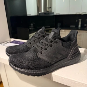 Adidas Ultraboost - Säljer ett par helsvarta Adidas Ultraboost sneakers med stickad ovandel och klassiska tre ränder på sidan. Skorna har snörning, platt sula och en riktigt bekväm boost-dämpning. Perfekta för dig som gillar stilrena och sportiga skor med modern känsla.