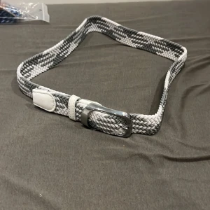 Vitt och grått flätat bälte - Snyggt flätat bälte i vitt och grått med silvrigt spänne. Bältet har detaljer i vitt konstläder vid ändarna och runt spännet. Passar perfekt till jeans eller chinos för en stilren look. Helt ny och oanvända