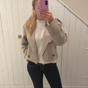 Beige croppad trenchcoat från Bershka - Säljer en beige trenchcoat från Bershka med stora knappar. Jackan har en loose fit och är perfekt för lager-på-lager. Klassisk krage och coola detaljer gör den superenkel att styla. Jackan är använd en gång bara. 