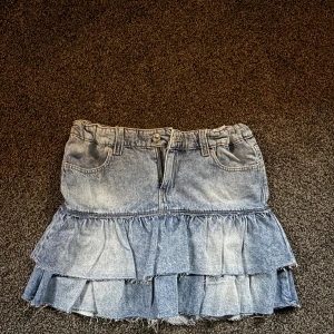 Blå volangkjol i denim från Gina Tricot - Supersöt blå jeanskjol från Gina Tricot med två lager volanger och råa kanter. Kjolen har klassiska fickor fram och bak, bälteshällor och stängs med knapp och dragkedja. Perfekt för dig som gillar en trendig och avslappnad stil.