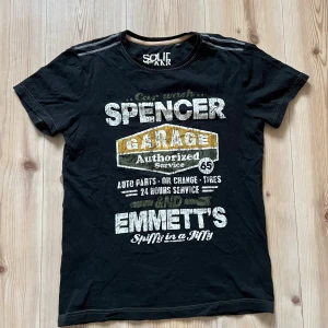 T-shirt Solid Jeans strl M - Cool svart t-shirt från Solid Jeans i storlek M med stort vintage-inspirerat tryck på bröstet i vitt, gult och grönt. Rund hals med beige kant och snygga sömdetaljer på axlarna. Perfekt för dig som gillar streetwear och grafiska prints.