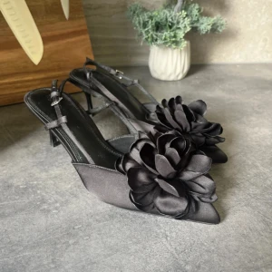 Svarta pumps med blomdetalj - Klackar från NA-KD, storlek 38, klack höjd 6/7 cm, inga skador på klack, skon eller sulan.                                                             Stilrena svarta pumps med spetsig tå och hög klack. Skorna har en stor dekorativ blomma framtill och smala remmar med spänne runt hälen. Tillverkade i satinliknande material som ger en lyxig känsla.