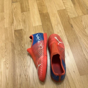 Puma Ultra röda fotbollsskor - Säljer ett par Puma Ultra fotbollsskor i rött och blått med vita detaljer och logga. Skorna har en strömlinjeformad design utan snörning och är tillverkade i syntetmaterial för bästa känsla på planen. Perfekta för dig som vill sticka ut med färg och stil på matchen.