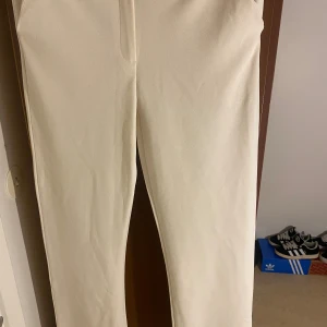 Beiga kostymbyxor från 157, storlek S - Snygga beiga kostymbyxor från 157 i storlek S. Byxorna har rak passform och är tillverkade i ett slätt, vävt material som ger en clean look. Perfekta för dig som gillar stilrena och minimalistiska plagg. Midjan är klassisk och byxorna har diskreta fickor framtill.