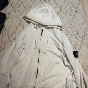 Vit hoodie från Stone Island, M - Vit hoodie från Stone Island med dragkedja och huva. Klassisk patch på vänster ärm och snörning i huvan. Tillverkad i mjuk bomull, perfekt för chill dagar.