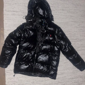 Svart glossy pufferjacka från Polo Ralph Lauren - Svart, glansig pufferjacka från Polo Ralph Lauren med huva och flera dragkedjor. Jackan har det klassiska röda Polo-logot broderat på bröstet och justerbara detaljer vid nederkant och huva. Perfekt för kalla dagar och har en riktigt snygg streetstil. Enda minus är att dragkedjan kan vara lite svår att sätta knäppa längstnere därpå de billiga priset, när man väll får i drag kedjan funkar den hur bra som helst. 1800 eller kom med bud
