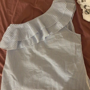 Randig one shoulder topp Vero Moda - Säljer en fräsch one shoulder topp från Vero Moda i storlek M. Toppen är vit och ljusblå med smala ränder och har en bred volang längs axeln. Materialet är lätt och känns som bomull, perfekt för varma dagar. Supersnygg till jeans eller kjol!