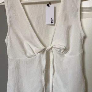 Vit omlottklänning från 157 - Superfin vit klänning från 157 med omlottknytning framtill och djup v-ringning. Ärmlös modell i ett lätt, strukturerat tyg som känns perfekt för varma dagar. Enkel och stilren design som passar till många tillfällen.