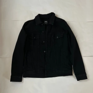 Svart jeansjacka från Zara - Svart jeansjacka från Zara med klassisk krage, knappar framtill och två bröstfickor med lock. Jackan har lång ärm och raka linjer för en clean look. Perfekt för dig som gillar stilrena och enkla plagg med edge.