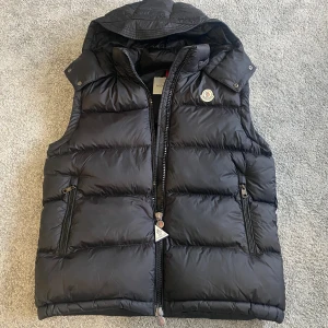 Svart Moncler väst med huva - Svart dunväst från Moncler med avtagbar huva, två dragkedjeförsedda fickor och ikonisk logga på bröstet. Västen har quiltad design och snygga detaljer på dragkedjan. Perfekt för lager-på-lager och streetstyle. De följer även me extra hängare till dragkedjan!