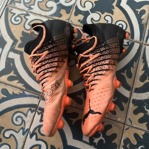 Puma Future fotbollsskor - Säljer ett par Puma Future fotbollsskor i orange och svart med coolt mönster och snörning. Skorna har högre strumpliknande skaft och greppig sula med dobbar för bästa fäste på planen. Perfekta för dig som vill sticka ut på matchen!