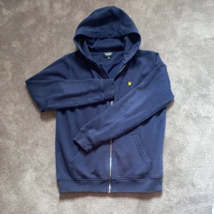 Svart zip hoodie från Lyle & Scott - Svart hoodie från Lyle & Scott med dragkedja framtill och klassisk gul logga på bröstet. Tröjan har långa ärmar, ribbade muddar och är tillverkad i mjuk bomull. Perfekt för en chill och stilren look. Hör av dig vid minsta fundering! Priset är prutbart.