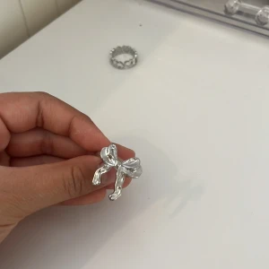 Silvrig ring med rosett - Superfin ring i silverfärg med en stor, glänsande rosett som detalj. Ringen har en modern och lekfull design som verkligen sticker ut. Perfekt accessoar för dig som vill ha något unikt och trendigt på fingret.