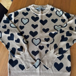 Brun guldig stickad tröja med svarta och glittriga hjärtan från Next - Superfin brun tröja från Next med svarta och guldiga hjärtan över hela. Guldiga trådar i hela tröjan. Tröjan har rund hals, långa ärmar och breda ribbade muddar. Perfekt för dig som gillar gulliga detaljer och vill sticka ut lite extra.