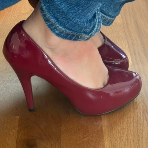 Vinröda pumps från Elissa - Snygga vinröda pumps från Elissa med hög klack och rund tå. Skorna har en glansig finish och är tillverkade i syntetmaterial. Perfekta för dig som vill sticka ut med en djup röd färg och klassisk siluett.