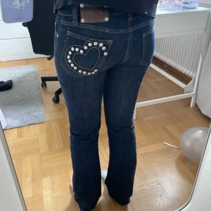 Moschino jeans med hjärtdetalj - Mörkblåa jeans från Moschino med snygga nitar i hjärtform på bakfickan. Klassisk femficksmodell med orange kontrastsömmar och bootcut-silhuett. Jeansen har låg midja och är tillverkade i kraftigt denimtyg. Säljer då de tyvärr har blivit för små. Använda ett fåtal gånger då de redan var lite små från början. 