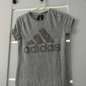 Grå Adidas t-shirt med logga - Snygg grå t-shirt från Adidas med stor svart logga framtill. Klassisk rund halsringning och korta ärmar. Perfekt för en sportig look eller till vardagsoutfiten. Mjuk och skön i bomullsmix.
