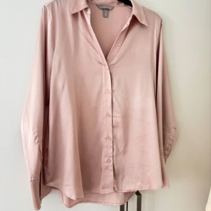 Ljusrosa silkesliknande skjorta H&M - Säljer en ljusrosa skjorta från H&M med silkesliknande material och klassisk krage. Skjortan har lång ärm, knappar framtill och en loose passform som ger en chill vibe. Perfekt för dig som gillar mjuka och glansiga plagg.