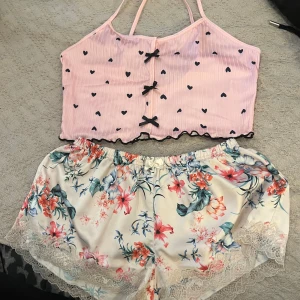 Rosa hjärtmönstrad linnetopp med rosetter - Supersöt rosa linnetopp med svarta hjärtan och tre små rosetter framtill. Toppen har tunna axelband, vågig kant nertill och är croppad. Perfekt för sommarens varma dagar eller som pyjamasöverdel.