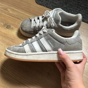 Grå Adidas Campus 00 sneakers strl 38 - Säljer ett par grå Adidas Campus sneakers i mocka med klassiska vita ränder på sidorna och vit snörning. Skorna har en platt gummisula i ljusbrunt och rund tå. Perfekt för dig som gillar retrostil och vill ha något bekvämt och snyggt till vardags. Typ nästan aldrig använda max 8-10 gånger 