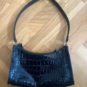 Svart axelväska i croco-imitation - Säljer en svart axelväska med croco-mönstrad yta och glansig finish. Väskan har en klassisk rektangulär form, dragkedja upptill och bred axelrem. Perfekt storlek för det viktigaste och ger en cool vibe till din outfit.