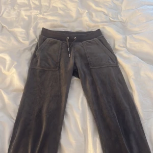 Gråa mjukisbyxor Juicy Couture XS - Mysiga gråa mjukisbyxor från Juicy Couture i velour med bred resår och snörning i midjan. Stora fickor fram och bak, bakfickorna har blingiga knappar. Broderad JC-logga på ena fickan framtill. Perfekta för chill dagar.