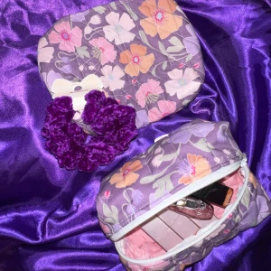 2 handgjorda necessärer och en virkad hårsnodd - Superfina handgjorda  necessärer i lila med blommigt mönster i pastellfärger, tillkommer med egenvirkad hårsnodd. Perfekt för smink eller småprylar.  Söt och färgglad accessoar som lyser upp din vardag.