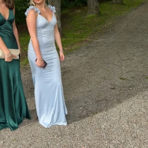 Ljusblå långklänning med volangaxlar - Supersnygg ljusblå långklänning i satinliknande material med volangdetaljer på axlarna och v-ringad urringning. Klänningen har smickrande passform, öppen rygg med knytband och är ärmlös. Perfekt för bal eller student! Använd en gång, så därav priset. PM mig för mer bilder!