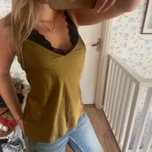 Olivgrön linnetopp med spetsdetalj - Säljer en olivgrön linnetopp med tunna axelband och snygg svart spets längs v-ringningen. Behövs bara strykas