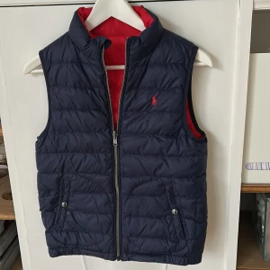 Vändbar dunväst från Polo Ralph Lauren - Vändbar dunväst från Polo Ralph Lauren med ena sidan marinblå och andra sidan röd. Västen har broderad logga på bröstet, två fickor med tryckknappar och dragkedja framtill. Perfekt för lager-på-lager nu när det börjar bli kallt och snygg att styla till jeans eller hoodie. Mycket bra skick, 9/10