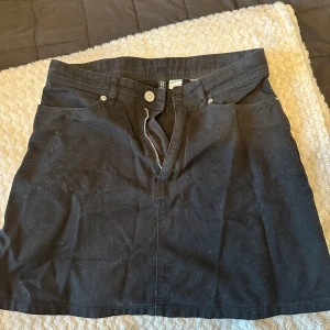 Svart kort jeanskjol från Divided - Svart kort jeanskjol från Divided med klassisk femficksdesign och dragkedja framtill. Snygg och enkel modell som passar till många olika stilar. Perfekt att matcha med en t-shirt eller hoodie för en avslappnad look.