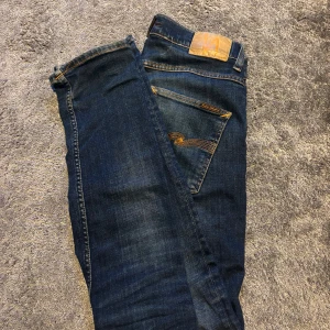 Mörkblå jeans från Nudie Jeans - Snygga mörkblå jeans från Nudie Jeans med klassiska orange sömmar och läderpatch bak i midjan. Jeansen har raka ben och femficksmodell, tillverkade i slitstarkt denimtyg. Perfekta för en avslappnad och stilren look. Modellen är Grim Tim