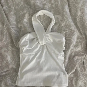 Vit ribbad halterneck topp - Säljer en vit ribbad halterneck topp med twistad detalj framtill. Toppen är ärmlös och har en figurnära passform som framhäver midjan. Perfekt att styla med jeans eller kjol för en trendig look.