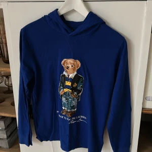 Blå tunn långärmad t shirt Polo Ralph Lauren - Blå hoodie från Polo Ralph Lauren med ikoniska Polo Bear-trycket framtill. Tröjan är en tunn långarmad t shirt med huva är gjord i mjuk bomull. Snygg och lätt att bära till flera olika outifts.