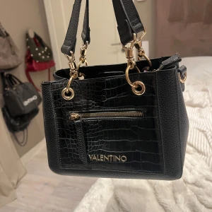 Svart handväska från Valentino - Snygg svart handväska från Valentino med croco-mönstrad framsida och guldfärgade detaljer. Väskan har både dragkedja och magnetlås, samt en praktisk ytterficka. Tillverkad i skinnimitation med struktur och bred axelrem. Slitna handtag, legat i garderoben ett tag