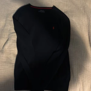 Marinblå stickad tröja Polo Ralph Lauren - Svart stickad tröja från Polo Ralph Lauren med klassisk röd broderad logga på bröstet. Tröjan har rund halsringning med röd detalj och långa ärmar. Perfekt för dig som gillar stilren och tidlös design.