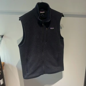 Mörkblå fleeceväst från Patagonia - Snygg mörkblå fleeceväst från Patagonia med hög krage och hel dragkedja framtill. Skick 8/10. Västen har två sidofickor med dragkedja och en bröstficka med dragkedja. Perfekt för lager-på-lager och outdoor vibes. Om det är något du undrar över så är det bara att fråga. 
