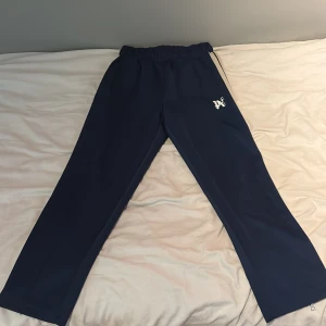 Palm Angels blå trackpants med dragkedja - Palm Angels trackpants i mörkblått med vit sidoremsa och svart detalj. Byxorna har elastisk midja med snörning, raka ben och dragkedja vid benslut. Ikonisk Palm Angels-logga på låret. Perfekta för en sportig och trendig look.