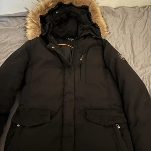 Svart dunjacka från Hollies med äkta päls - Säljer en svart parkas från Hollies med huva och äkta päls. Jackan har flera fickor med dragkedja och tryckknappar, samt en snygg logga på ärmen. Perfekt för kalla dagar med sin varma och rejäla känsla.