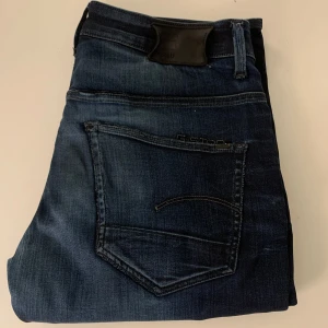 Mörkblå jeans från G-Star RAW, W31 L32 - Snygga mörkblå jeans från G-Star RAW med klassisk femficksdesign och diskreta slitningar. Jeansen har raka ben och en cool tvättad look. Perfekta för dig som gillar en stilren men ändå trendig vibe.