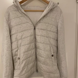 Ljusgrå quiltad cardigan med huva - Snygg ljusgrå cardigan med quiltad framsida och mjuka ärmar. Jackan har huva, dragkedja hela vägen och två fickor med knappstängning. Perfekt för dig som vill ha en stilren och bekväm jacka till vardags.