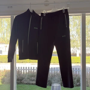 Svart ziptröja från BOSS med gröna detaljer - Svart ziptröja från BOSS med gröna detaljer och logga på bröstet. Tröjan har hel dragkedja framtill, två fickor med dragkedja och lång ärm. Materialet är mjukt och sportigt, perfekt för en chill och stilren look. EJ FRAKT MÖTS BARA UPP I VÄSTRA GÖTALAND.