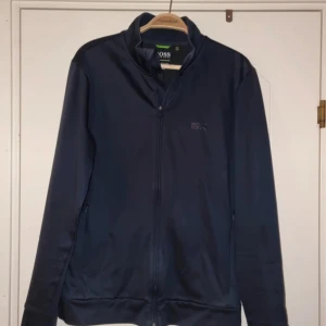 Marinblå ziptröja från Hugo Boss - Snygg marinblå ziptröja från Hugo Boss med reflex logotryck på ryggen. Tröjan har lång ärm, hög krage och dragkedja framtill. Slim fit-modell i mjukt material, perfekt för en stilren och sportig look.                                       Mycket fint skick. Storlek M.