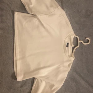 Vit croppad topp med kort ärm - Enkel och stilren vit croppad topp med korta ärmar och rund halsringning. Toppen har en boxig passform och är gjord i mjukt bomullstyg, perfekt för en avslappnad look.