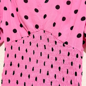 Rosa prickig kortklänning med volang - Superfin rosa kortklänning med svarta prickar och v-ringad hals. Klänningen har puffiga kortärmar med rynkade muddar och en smockad midja som ger en slim passform. Volangdetalj längst ner för extra cute vibe. Perfekt för dig som gillar färg och mönster! Materialet är 100% Rayon.