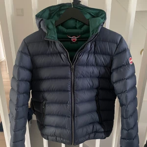 Mörkblå Colmar pufferjacka med huva - Säljer en mörkblå pufferjacka från Colmar med grön insida och huva. Jackan har dragkedja framtill, två fickor med dragkedja och klassisk Colmar-logga på ärmen. Perfekt för kyliga dagar och riktigt snygg passform. En liten dov fläck finns på höger arm. Har inte provat tvätta den. Finns att se på sista bilden.